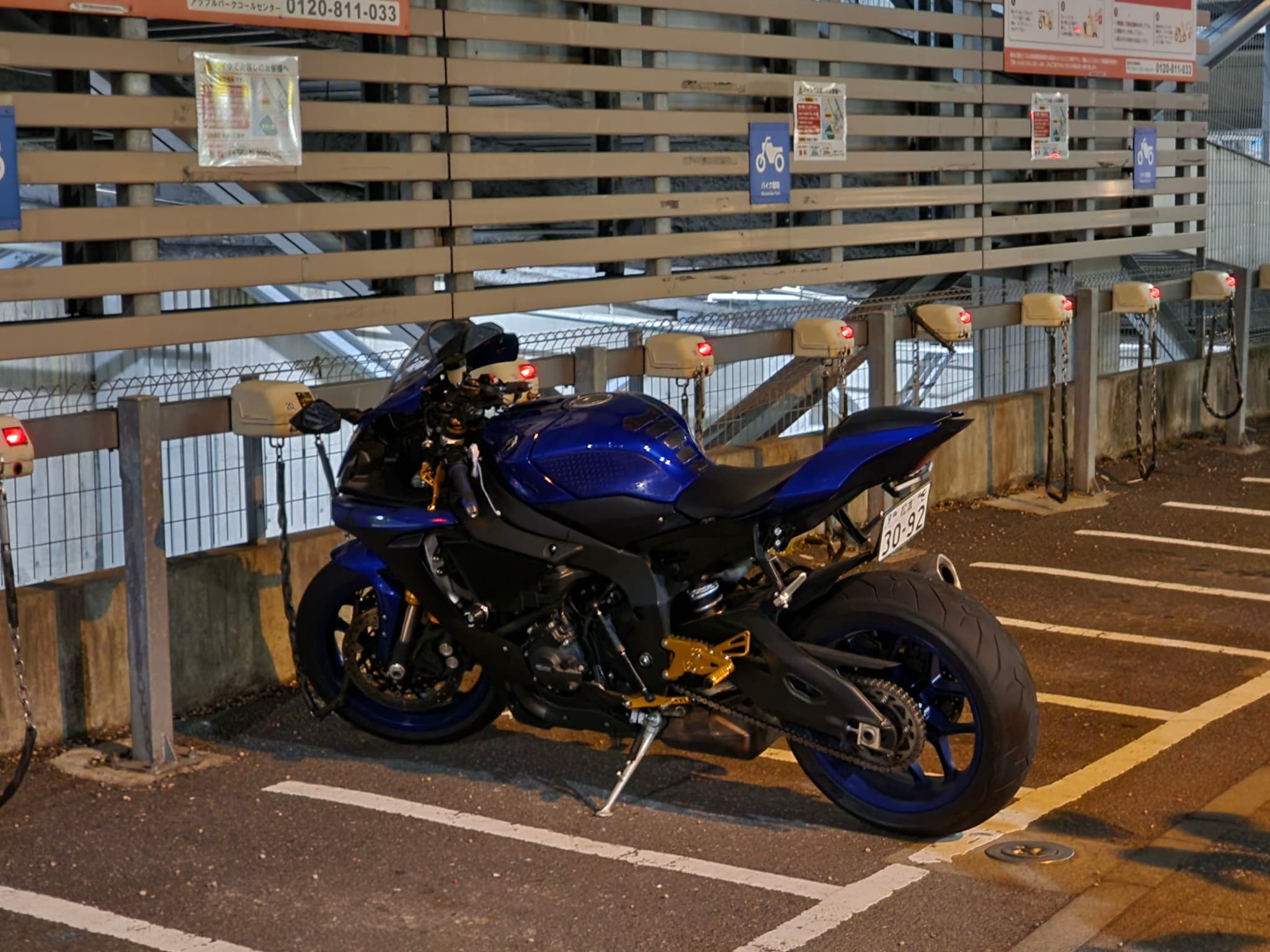 A blue motorcycle is parked in a designated parking space near a fence with various signs attached to it