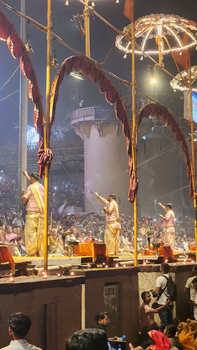 A group of people performing the Ganga Aarti ceremony at Dashashwamedh Ghat in Varanasi, India, with priests dressed in traditional attire holding ritual items The scene is illuminated by lights and surrounded by a crowd of onlookers